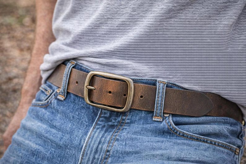 May include: A brown leather belt with a distressed finish and a rectangular brass-toned metal buckle. The belt is threaded through the belt loops of blue denim jeans, worn with a light gray textured t-shirt tucked into the waistband.