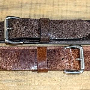 May include: Two brown leather belts with silver-colored buckles. The top belt is a darker shade of brown, while the bottom belt is a lighter shade. Both belts have a textured, distressed appearance.