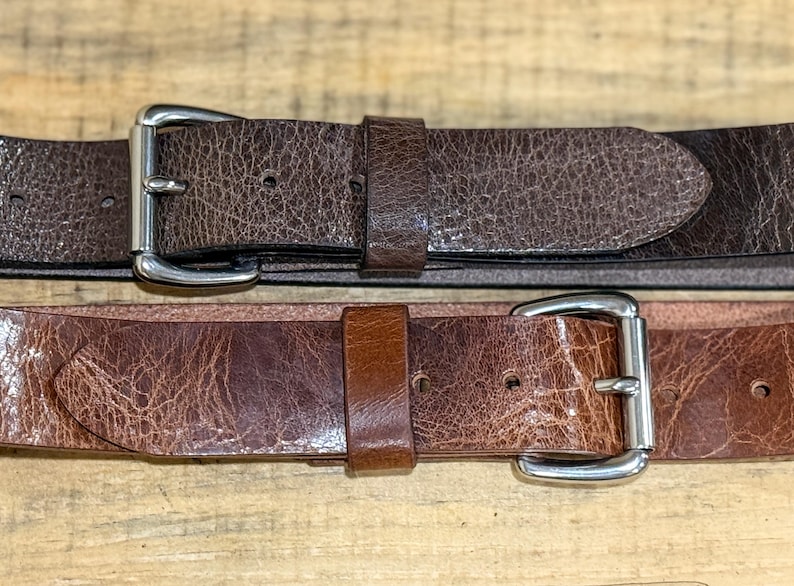 May include: Two brown leather belts with silver-colored buckles. The top belt is a darker shade of brown, while the bottom belt is a lighter shade. Both belts have a textured, distressed appearance.