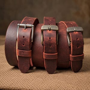 May include: Three rolled belts made of distressed reddish-brown leather. Each belt features a distinct metal buckle, including one silver-toned rectangular buckle and two antique brass-toned buckles, with matching leather keepers and holes.