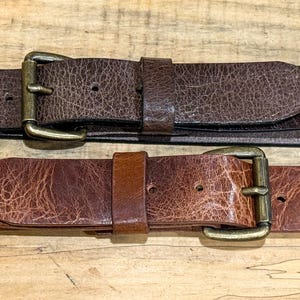 May include: Two brown leather belts with brass buckles are displayed on a wooden surface. The belts have a textured appearance, with one being a darker shade of brown than the other. The buckles are rectangular and have a classic design.