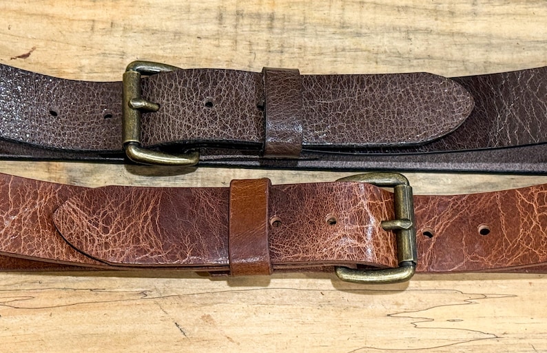 May include: Two brown leather belts with brass buckles are displayed on a wooden surface. The belts have a textured appearance, with one being a darker shade of brown than the other. The buckles are rectangular and have a classic design.