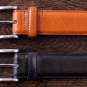 May include: Two belts, one in tan and one in black, each featuring a smooth leather-like texture, matching stitching along the edges, a single leather-like keeper loop, and a rectangular silver-toned metal buckle with a single prong closure.