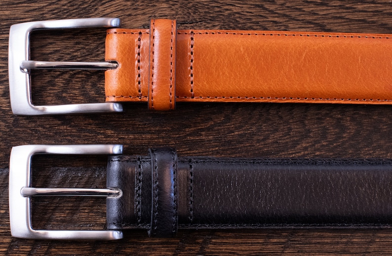 May include: Two belts, one in tan and one in black, each featuring a smooth leather-like texture, matching stitching along the edges, a single leather-like keeper loop, and a rectangular silver-toned metal buckle with a single prong closure.