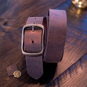 A dark brown leather belt with a brass-colored buckle is coiled on a dark wooden surface, with two small leather tags nearby.