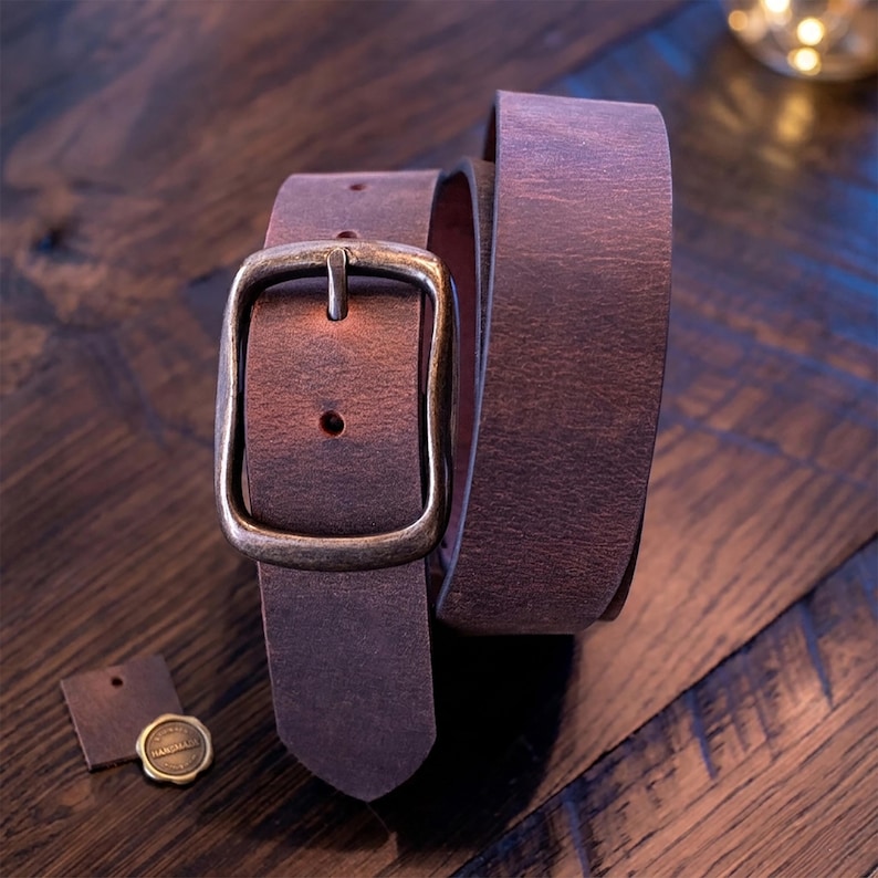 A dark brown leather belt with a brass-colored buckle is coiled on a dark wooden surface, with two small leather tags nearby.