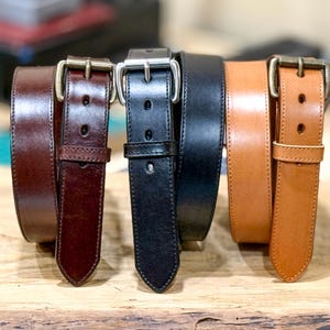 May include: Four leather belts in shades of brown and black are displayed. The belts have metal buckles and keepers. The belts are made of smooth leather and appear to be of high quality. The belts are displayed on a wooden surface.
