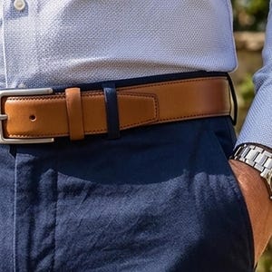 May include: A light blue textured long-sleeve shirt tucked into navy blue trousers, featuring a tan leather belt with a silver-toned metal buckle and a silver-toned metal wristwatch with a black dial worn on the left wrist.