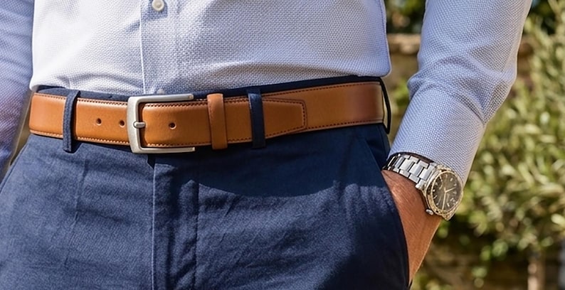May include: A light blue textured long-sleeve shirt tucked into navy blue trousers, featuring a tan leather belt with a silver-toned metal buckle and a silver-toned metal wristwatch with a black dial worn on the left wrist.