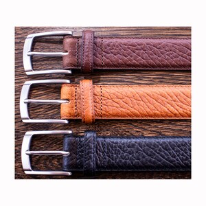 Pode incluir: Tr&ecirc;s cintos com superf&iacute;cies texturizadas em castanho escuro, castanho claro e preto. Cada cinto possui uma fivela de metal prateado com um pino simples e um passador de couro a condizer. As tiras t&ecirc;m costuras vis&iacute;veis ao longo das bordas.