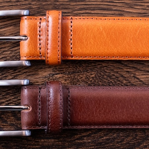 May include: Two leather belts with silver-toned metal buckles. The top belt is a light tan color, and the bottom belt is a dark brown color. Both belts feature matching leather keepers and visible stitching along the edges of the straps.