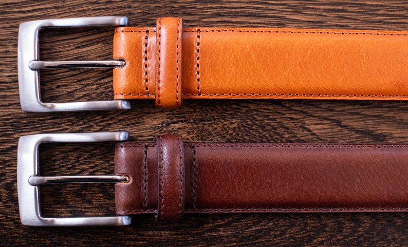 May include: Two leather belts with silver-toned metal buckles. The top belt is a light tan color, and the bottom belt is a dark brown color. Both belts feature matching leather keepers and visible stitching along the edges of the straps.
