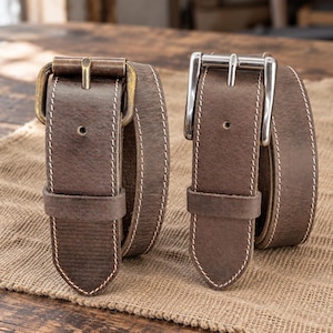 May include: Two brown leather belts with white contrast stitching. The left belt features an antiqued brass-tone metal buckle, while the right belt has a polished silver-tone metal buckle. Both belts are rolled and stand upright on their ends.