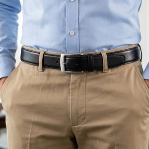 May include: A black leather belt with a silver-toned metal buckle worn with khaki trousers and a light blue button-down shirt. The belt features a smooth finish, a single prong, and a matching leather keeper loop, secured through the belt loops.