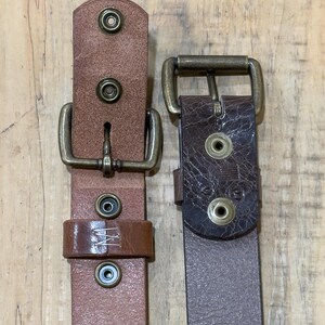 May include: Two leather belts with brass buckles and rivets. One belt is a light brown color, and the other is a darker brown. The belts are displayed against a wooden background.