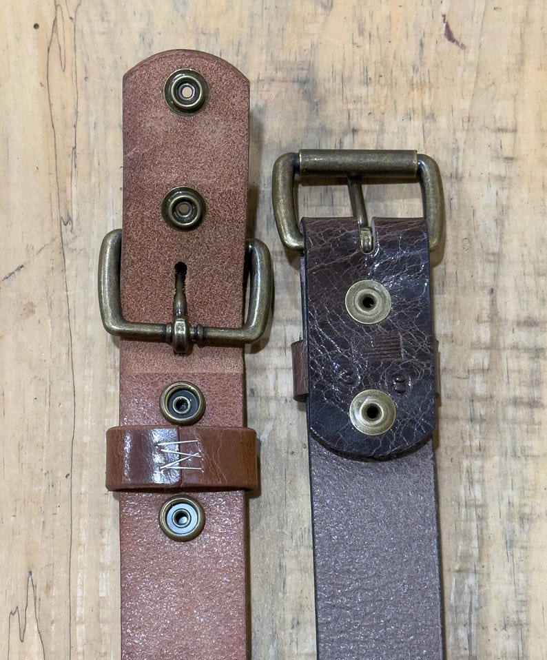 May include: Two leather belts with brass buckles and rivets. One belt is a light brown color, and the other is a darker brown. The belts are displayed against a wooden background.