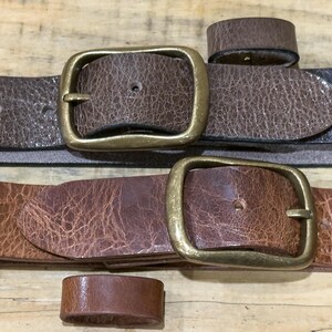 May include: Close-up of two brown leather belts with brass buckles and matching belt loops. The belts have a textured appearance and varying shades of brown. The buckles are rectangular and have a vintage look.