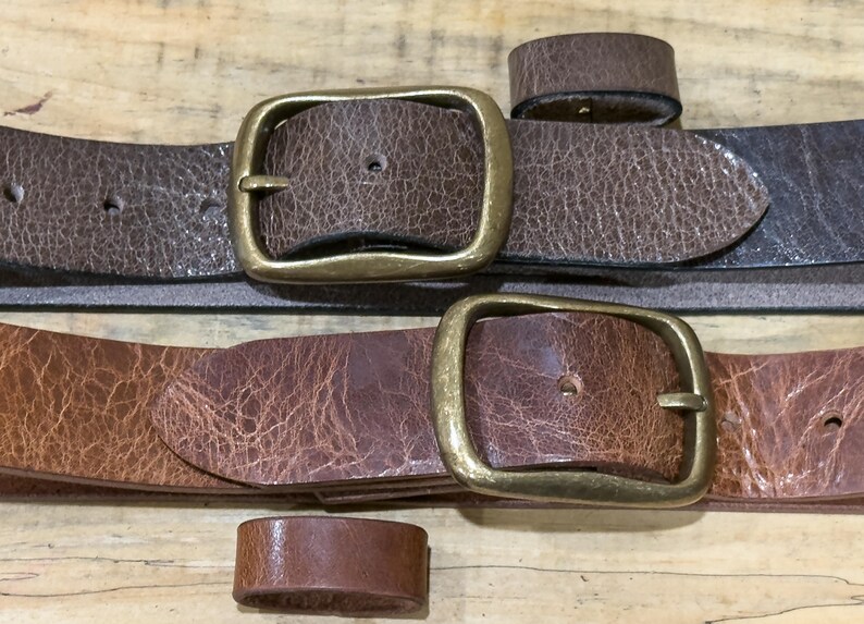 May include: Close-up of two brown leather belts with brass buckles and matching belt loops. The belts have a textured appearance and varying shades of brown. The buckles are rectangular and have a vintage look.