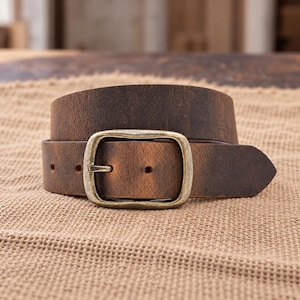 May include: A dark brown leather belt with a distressed finish and a rectangular antique brass-toned metal buckle. The belt features a pointed tip and visible adjustment holes, showcasing a weathered texture across the strap surface.