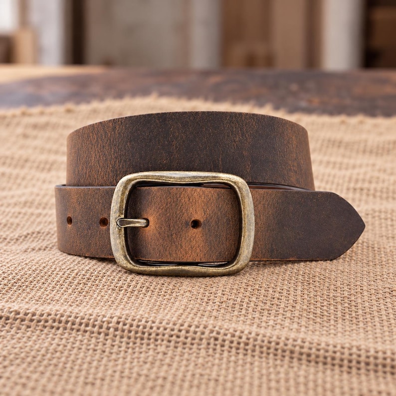 May include: A dark brown leather belt with a distressed finish and a rectangular antique brass-toned metal buckle. The belt features a pointed tip and visible adjustment holes, showcasing a weathered texture across the strap surface.