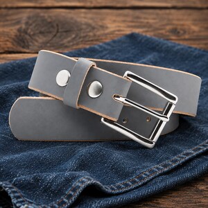 May include: A grey leather belt featuring a polished silver-tone metal buckle and two matching silver-tone metal rivets securing the belt loop. The belt has a smooth matte finish and a rounded tip, showcasing a clean and functional design.