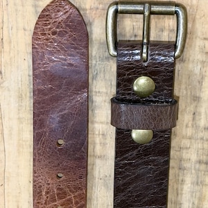 May include: Close-up of a brown leather belt with a brass buckle. The belt has a distressed texture and visible stitching. The buckle is rectangular with a single prong. The belt is approximately 1.5 inches wide.