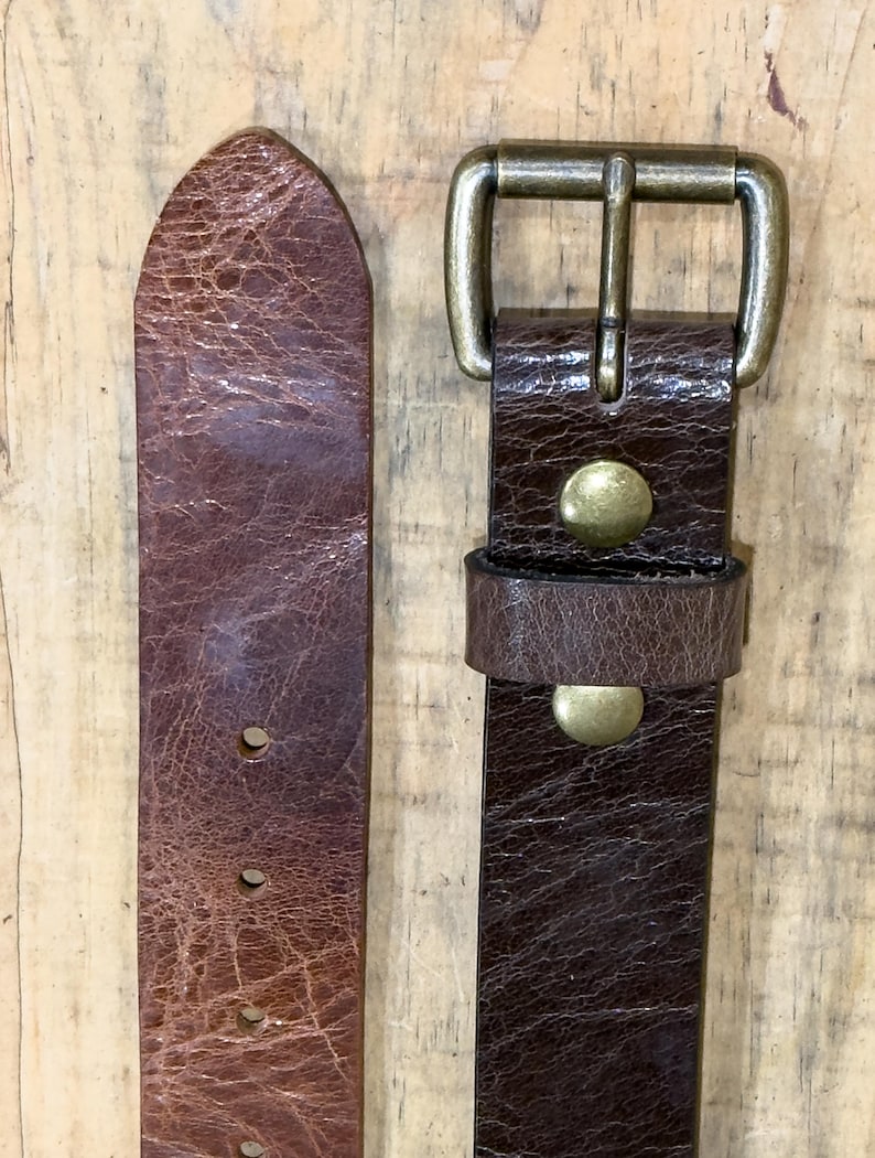 May include: Close-up of a brown leather belt with a brass buckle. The belt has a distressed texture and visible stitching. The buckle is rectangular with a single prong. The belt is approximately 1.5 inches wide.