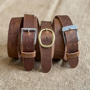 May include: Three brown leather belts coiled together, each featuring a distinct metal buckle. The left belt has a rectangular silver buckle, the center belt has an oval brass buckle, and the right belt has a rectangular brushed metal buckle.