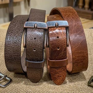 May include: Two leather belts with metal buckles. One belt is dark brown, the other is a lighter shade of brown. The belts are displayed on a burlap surface, showcasing their texture and craftsmanship. The buckles have a rustic, aged appearance.
