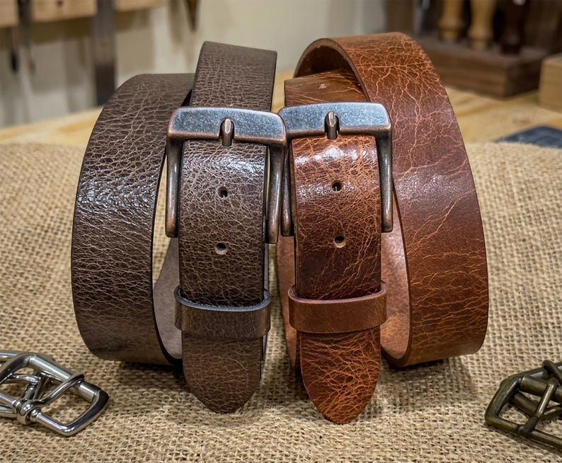 May include: Two leather belts with metal buckles. One belt is dark brown, the other is a lighter shade of brown. The belts are displayed on a burlap surface, showcasing their texture and craftsmanship. The buckles have a rustic, aged appearance.