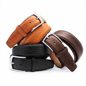 Pode incluir: Uma pilha de quatro cintos de couro com uma textura granulada distinta. A cole&ccedil;&atilde;o inclui um cinto castanho-claro, um castanho-escuro e dois pretos, cada um equipado com uma fivela de metal prateado e um passador de couro a condizer.