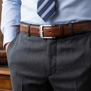 May include: A light blue button-down shirt with a navy and white striped tie, a brown leather belt with a silver-toned buckle, and dark gray trousers. A silver wristwatch with a brown leather strap is visible on the left wrist.