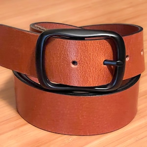 A tan leather belt with a black metal buckle. The belt is folded in half and the buckle is visible in the center of the image.