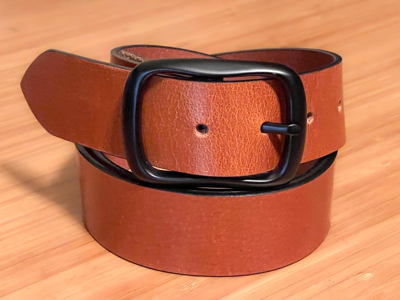 A tan leather belt with a black metal buckle. The belt is folded in half and the buckle is visible in the center of the image.