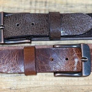 May include: Two leather belts with metal buckles. The top belt is a dark brown with a textured surface, and the bottom belt is a lighter brown with a similar texture. Both belts have a classic design.