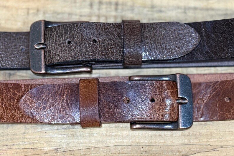 May include: Two leather belts with metal buckles. The top belt is a dark brown with a textured surface, and the bottom belt is a lighter brown with a similar texture. Both belts have a classic design.
