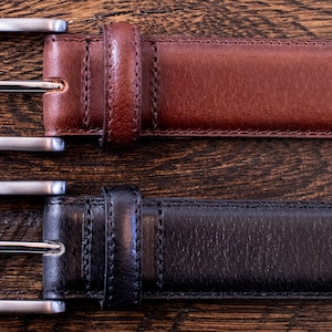 May include: Two leather belts, one in brown and one in black, each featuring a silver-toned metal buckle with a rectangular frame and a single prong. The belts have visible stitching along the edges and a matching leather keeper loop near the buckle.