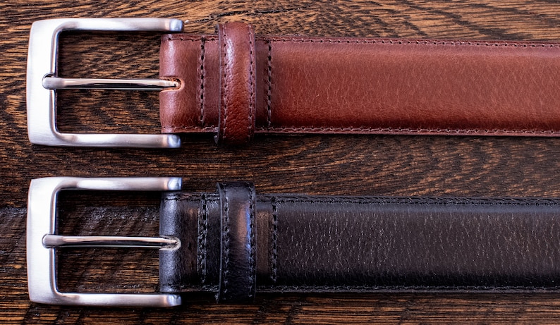 May include: Two leather belts, one in brown and one in black, each featuring a silver-toned metal buckle with a rectangular frame and a single prong. The belts have visible stitching along the edges and a matching leather keeper loop near the buckle.