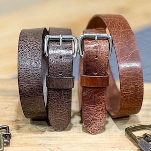May include: Two leather belts with silver-colored buckles. One belt is a dark brown color, and the other is a lighter brown. The belts are displayed on a wooden surface. The leather has a textured appearance.