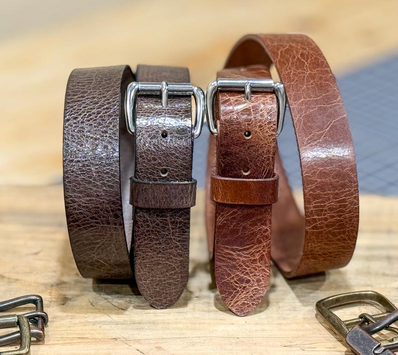 May include: Two leather belts with silver-colored buckles. One belt is a dark brown color, and the other is a lighter brown. The belts are displayed on a wooden surface. The leather has a textured appearance.