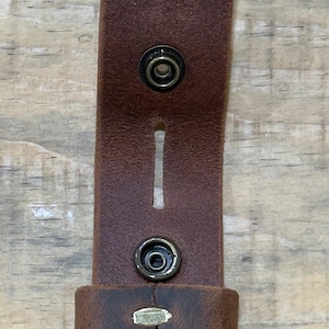 May include: A brown leather strap featuring four circular metal snap fasteners arranged in a vertical line. The strap includes a rectangular cutout between the second and third snaps and a leather loop positioned near the bottom snap.