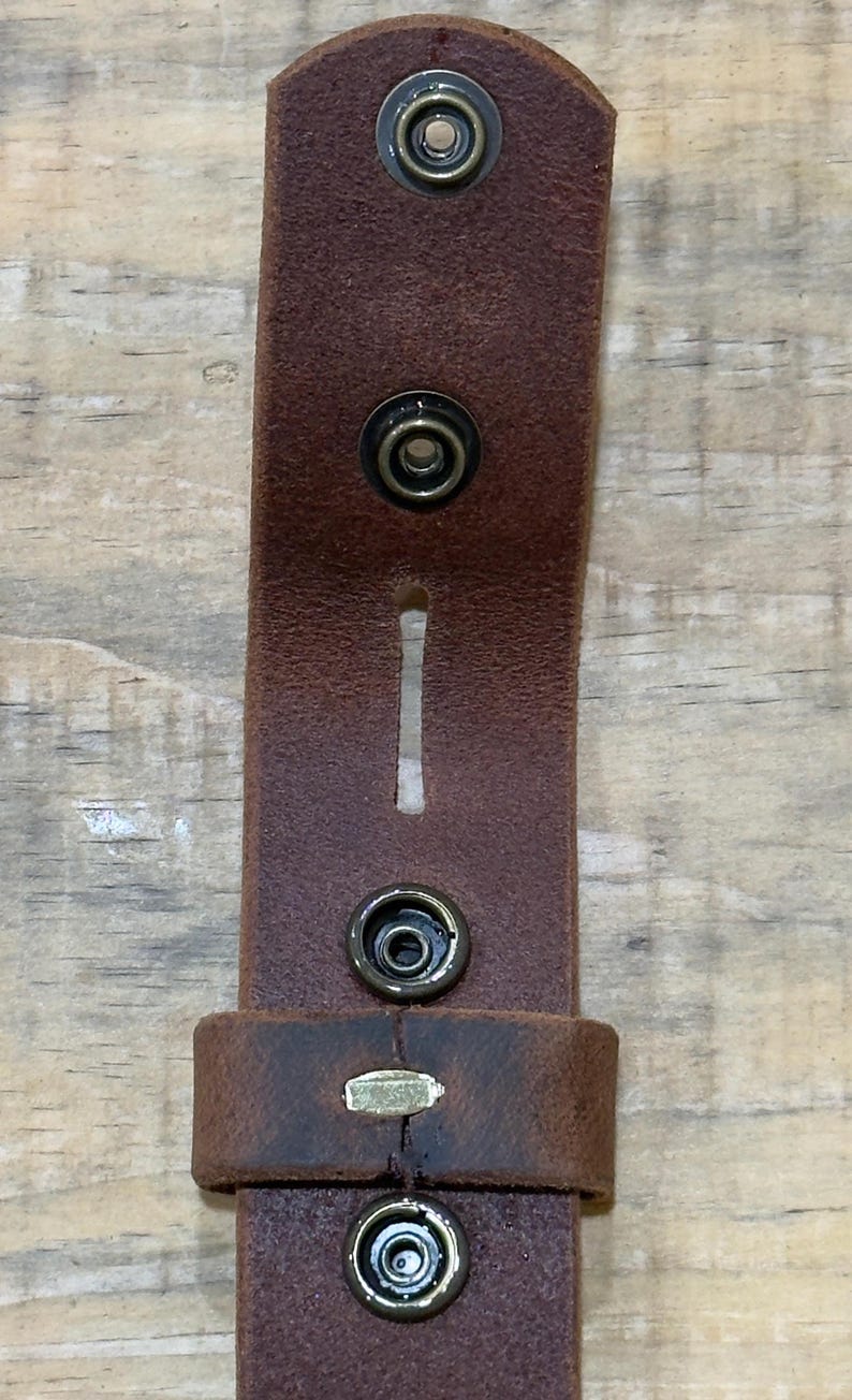 May include: A brown leather strap featuring four circular metal snap fasteners arranged in a vertical line. The strap includes a rectangular cutout between the second and third snaps and a leather loop positioned near the bottom snap.