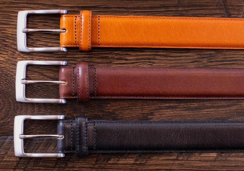 May include: Three belts arranged horizontally, each featuring a smooth finish and a silver-toned metal buckle. The top belt is tan, the middle belt is dark brown, and the bottom belt is black. Each belt includes a matching leather keeper loop.