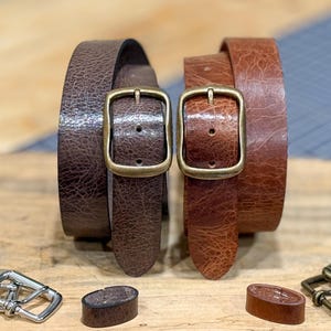May include: Two leather belts in shades of brown, each with a rectangular brass buckle. The belts are rolled up, showcasing their texture. Additional belt hardware is visible at the bottom. The belts appear to be approximately 1.25 inches wide.