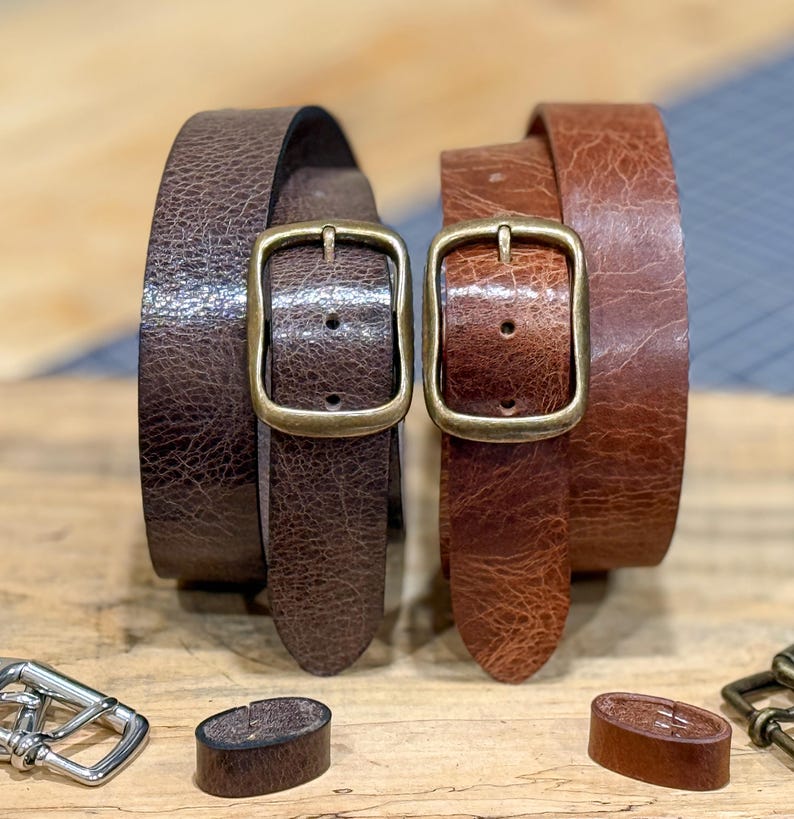 May include: Two leather belts in shades of brown, each with a rectangular brass buckle. The belts are rolled up, showcasing their texture. Additional belt hardware is visible at the bottom. The belts appear to be approximately 1.25 inches wide.