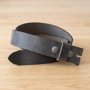 May include: A dark charcoal gray leather belt featuring a textured surface, a pointed tip, and two circular metallic studs securing the strap. The belt is coiled into a circular shape, showcasing the matte finish of the leather and the hardware.
