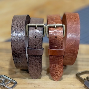 May include: Two leather belts in shades of brown, with brass-colored buckles. The belts have a textured appearance, and are displayed on a wooden surface. The belt on the left is a darker brown, while the belt on the right is a lighter, reddish-brown.
