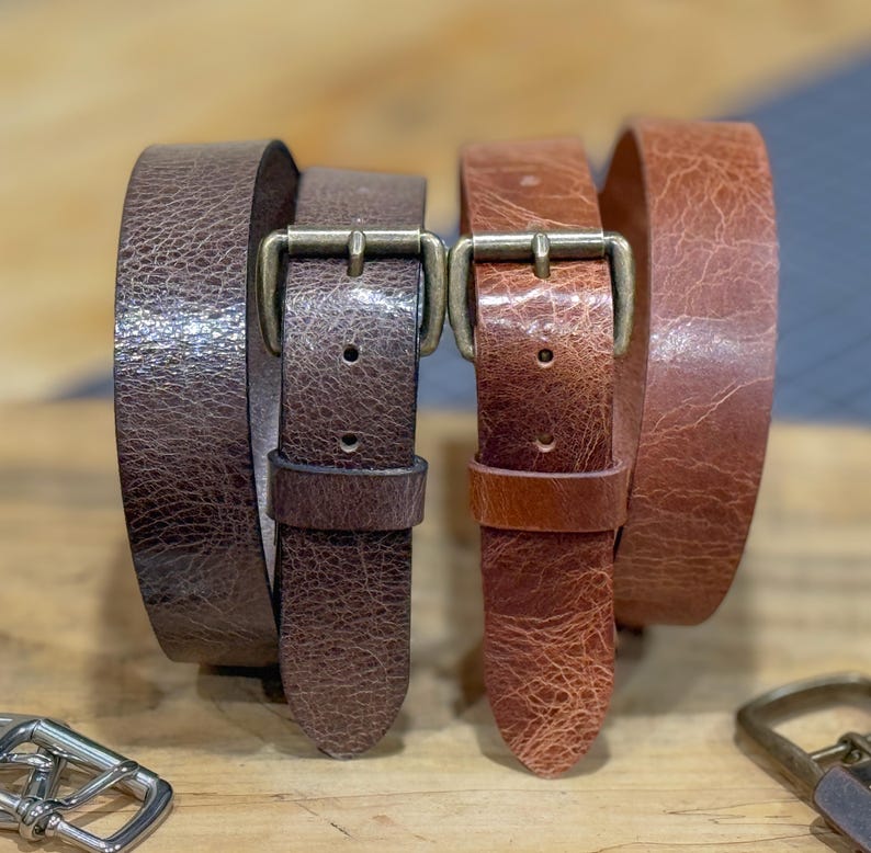 May include: Two leather belts in shades of brown, with brass-colored buckles. The belts have a textured appearance, and are displayed on a wooden surface. The belt on the left is a darker brown, while the belt on the right is a lighter, reddish-brown.