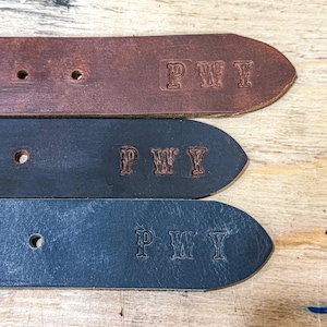 May include: Three leather belt ends in brown, dark gray, and black, each featuring the embossed letters PWY. The belts have visible adjustment holes and a smooth texture, showcasing the stamped personalization on the tapered ends.