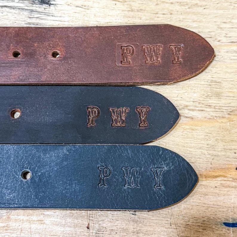 May include: Three leather belt ends in brown, dark gray, and black, each featuring the embossed letters PWY. The belts have visible adjustment holes and a smooth texture, showcasing the stamped personalization on the tapered ends.